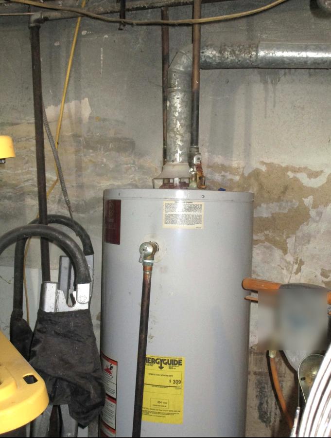 Old gas water heater with a corroded flue against a water-stained basement wall before replacement
