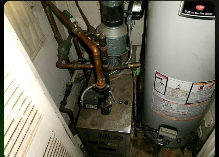 Old boiler in a cramped closet with tangled copper piping and a rusting galvanized flue before replacement